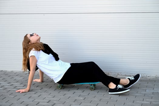 A person with shoulder-length light brown hair, wearing a white t-shirt and a black scarf, is holding a bright blue skateboard on their shoulder, standing against a gray, horizontally-paneled wall photo