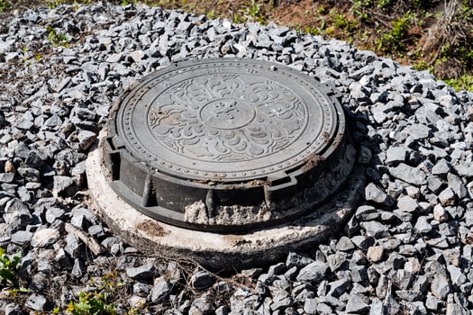 Ornate metal manhole cover surrounded by gravel stones in outdoor urban setting, showcasing decorative design. A functional utility cover providing access to underground infrastructure photo