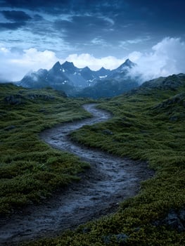 A winding path leads through a lush green landscape towards majestic mountains under a dramatic sky photo