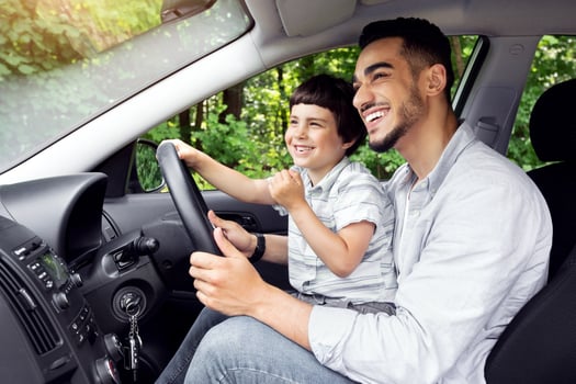 Young Arab Father Giving Driving Lessons To His Little Son, Happy Preteen Boy Sitting On Dad's Lap In Modern Car And Holding Steering Wheel, Enjoying Road Trip And Spending Time Together, Side View photo