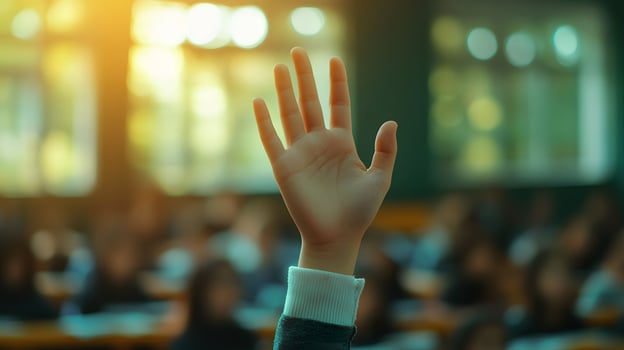 White-sleeved hand raises in classroom environment, representing participation and volunteering against blurred background with warm lighting and educational context. responsive photo