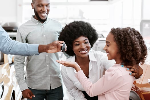 Positive auto salesman giving car key to cute black girl and her parents at dealership store. Cheerful African American family shopping for new vehicle at automotive showroom photo