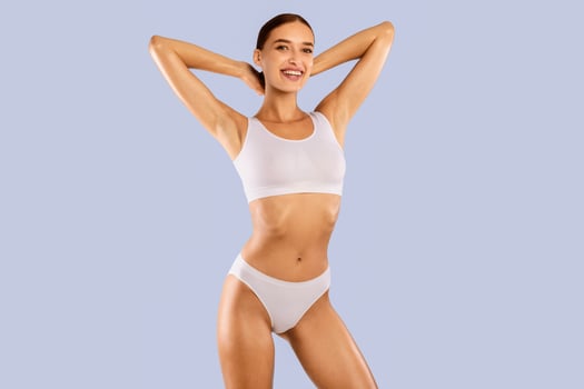 Portrait of happy smiling fit woman wearing lingerie posing isolated om blue studio background, raising arms up. Female with flat belly in white underwear top bra and panties showing smooth armpits photo
