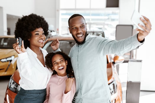 Joyful Afro family taking selfie with car key after buying new vehicle at auto dealership. Excited black clients making photo of themselves near modern automobile at showroom