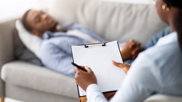 Stress and psychological trauma. Psychologist working with depressed black man at office, mockup on clipboard. African American male patient lying on couch while having session with therapist photo