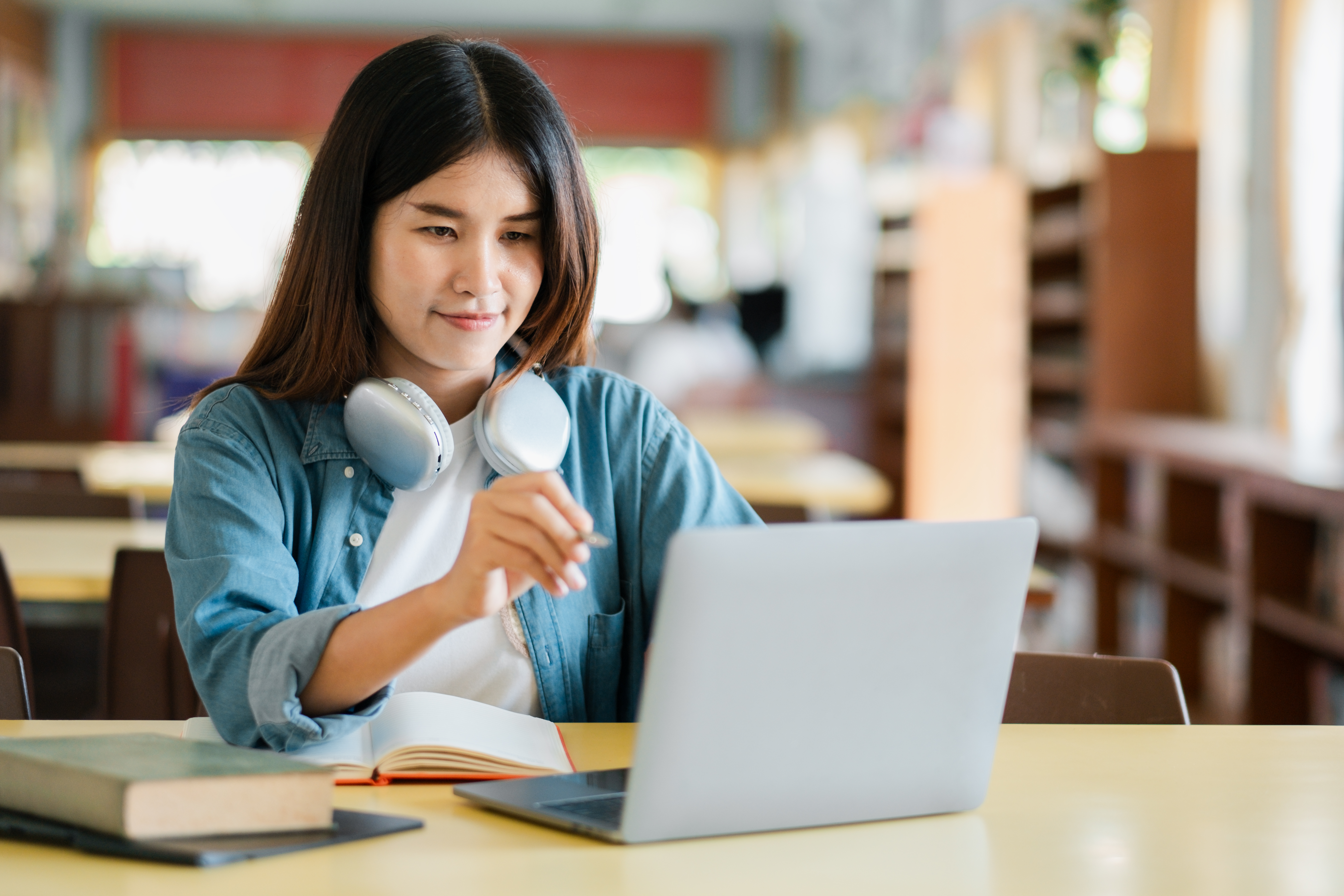 A young woman in a library using a laptop, wearing headphones, surrounded by books, focused on studying and research. photo