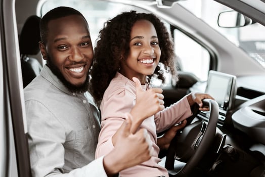 Positive black man and his cute daughter sitting on driver seat of new car, showing thumbs up at auto dealership. Friendly African American family test driving vehicle together at showroom photo