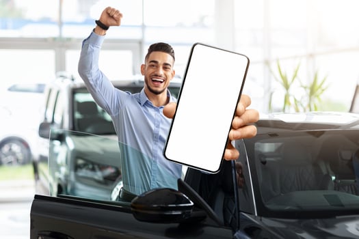 Emotional middle eastern guy choosing new car, standing by brand new black automobile, showing modern cell phone with white blank screen and raissing hand up, using auto dealership app, mockup photo