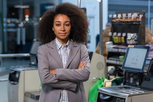 A confident supermarket cashier manager with curly hair in business attire standing with arms crossed at a checkout counter. photo