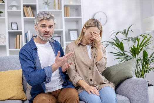 Middle-aged couple having an argument at home, with the man gesturing and the woman covering her face in frustration on the sofa. photo