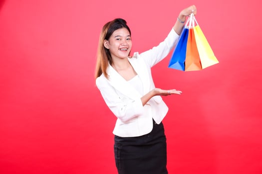 A woman holds three colorful paper shopping bags with her arm raised, while the other hand is showing an open palm, isolated on a red background photo