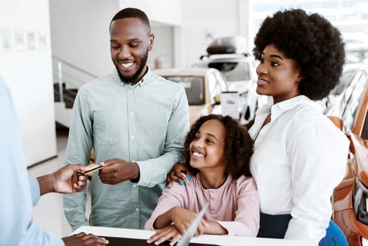 Cheerful black family paying for new car with credit card, making deal with salesman at auto showroom. African American clients purchasing automobile at modern dealership photo
