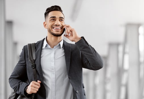 Smiling Middle Eastern Man Talking On Mobile Phone While Walking At Airport Terminal, Happy Young Arab Businessman Carrying Hand Luggage And Enjoying Pleasant Conversation On Cellphone, Copy Space photo