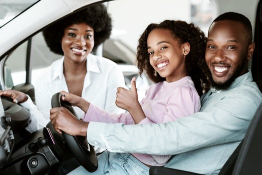 Cute black girl with her parents recommending auto dealership, sitting inside new car, showing thumb up gesture and smiling at automobile showroom store. Successful test drive photo