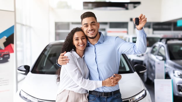 Happy young family buying new car in luxury showroom, standing buy brand new white auto, embracing and showing automatic key, copy space. Cheerful couple bought nice auto, car store interior photo
