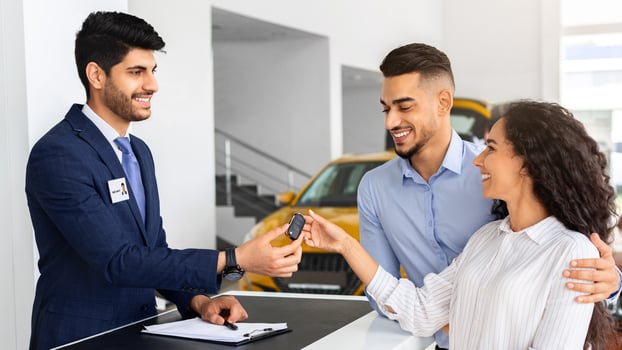 Happy middle-eastern couple buying car in huge showroom, taking automobile key from young handsome salesman, side view. cheerful arab man and woman purchasing beautiful auto, hugging and smiling photo
