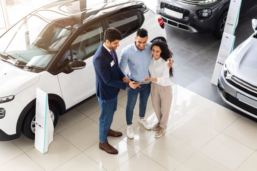 Top view of cheerful middle-eastern young couple having conversation with good-looking sales assistant at auto showroom, checking papers or car specification, full length photo