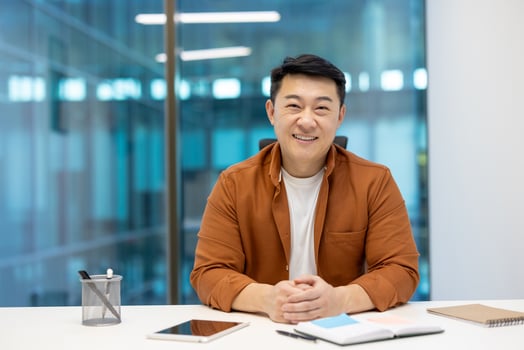 Asian businessman smiling confidently while sitting at office desk with tablet and notebook visible. Represents professionalism, positivity, and success in business setting. photo