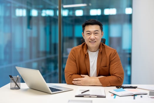 Asian businessman seated at desk with laptop, documents nearby. Casual attire, genuine smile conveying professionalism, sincerity. Modern office setting reflecting successful corporate atmosphere. photo
