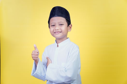 An asian boy in traditional Muslim attire poses giving a thumbs up against a yellow background, the concept of the Ramadan Kareem celebration. photo