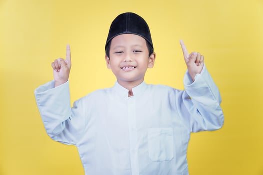 An asian boy in traditional Muslim dress poses pointing upwards with both hands against a yellow background, the concept of the ramdan kareem celebration photo