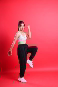 A woman in a white top and black pants with white shoes is striking a pose with one arm raised and one leg lifted, isolated on a red background photo