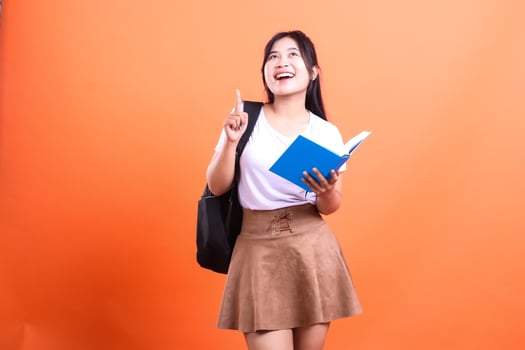 A young woman with a book and backpack points upward with an orange background behind her student photo