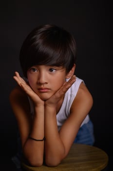 A young Asian boy stands in a studio, dressed in a white tank top and jeans, his silhouette contrasting sharply against the black background. photo