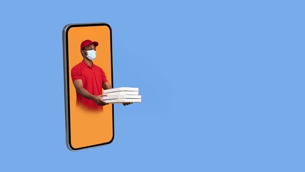Black Deliveryman In Cellphone Giving Pizza Boxes To The Side Wearing Protective Face Mask Delivering Restaurant Food Over Yellow Studio Background. Panorama With Copy Space, Collage. Delivery App photo