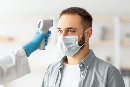 Doctor measuring young guy's temperature with electronic thermometer at home. Caucasian man in face mask checking his health before coronavirus immunization. Vaccination against covid concept photo