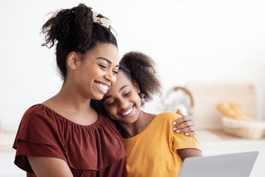 Cute black mother and kid using laptop together, sitting at kitchen table with homemade pastry, hugging and looking at computer screen, spending time together, watching cartoon, copy space photo