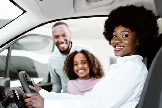 Test drive. Happy black woman sitting in driver seat of new car, choosing vehicle with husband and daughter at auto dealership. Afro family checking automobile before purchase or rental photo