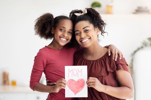 Cute black girl teenager greeting her beautiful mom with International Mothers Day, happy african american mother and daughter holding gift card and smiling at camera, kitchen interior, copy space photo