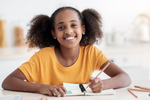 Cheerful inteligent african american teen girl in casual doing homework, sitting at cozy kitchen at home, taking notes or making plan, using pencils and notepad, smiling at camera, closeup portrait photo