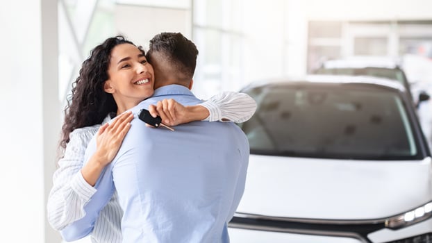 Ecstatic young curly woman with key from brand new car hugging her loving husband and smiling, feeling thankful and happy, standing by luxury auto, showroom interior, panorama with copy space photo