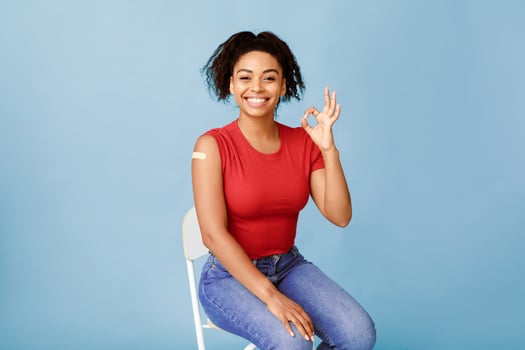 I recommend flu vaccine. Joyful young African American lady with vaccinated arm after flu injection showing okay gesture and smiling at camera on blue studio background. Antiviral vaccination campaign photo