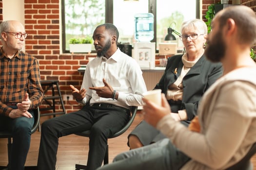 Young black guy sharing his mental health struggles with aa group community in brick wall office. African american man sitting in circle and talking about his recovery progress during therapy session. photo