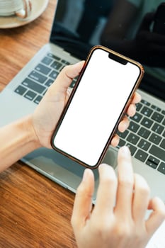 Close-up of a hand holding a smartphone with a blank screen above a laptop on a wooden desk. photo