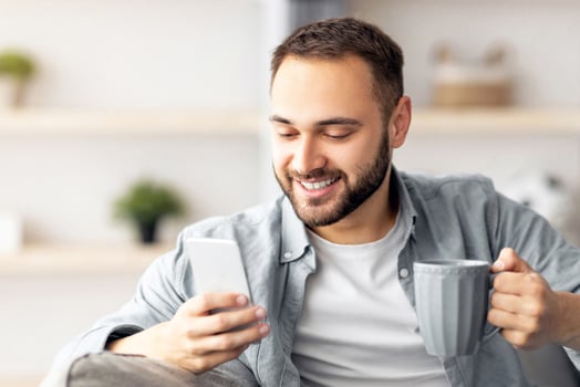 Positive millennial guy holding modern smartphone and cup of coffee indoors, chatting with friends, sending email, browsing internet or watching entertainment . Online life concept photo