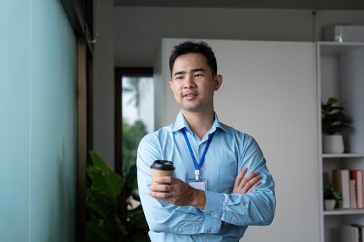 Confident Asian businessman holding a coffee cup, standing with arms crossed by a window in a modern office space. photo