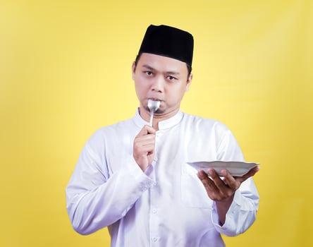 An Asian Muslim man in traditional dress bites into a spoon while holding a plate photo