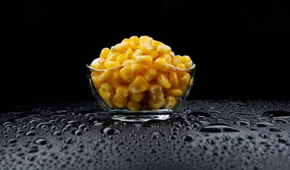 A transparent glass cup full of canned corn kernels on a black background with water drops.Canned corn kernels... photo