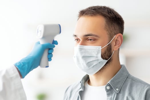 Doctor in rubber gloves measuring young guy's temperature with infrared thermometer indoors. Millennial man in medical mask checking health before coronavirus vaccine shot. Immunization against covid photo