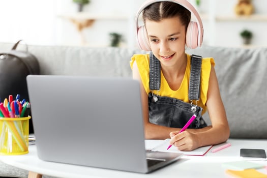 Online Education Concept. Smiling young teenage girl in headphones sitting at desk, using laptop writing or drawing in her paper notebook with colorful marker. Schoolgirl doing creative homework photo