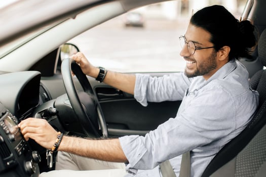 Cheerful long-haired stylish arab guy with fasten seat belt turning on music in car, happy middle-eastern young man having trip by auto, regulating music volume on automobile dashboard, copy space photo