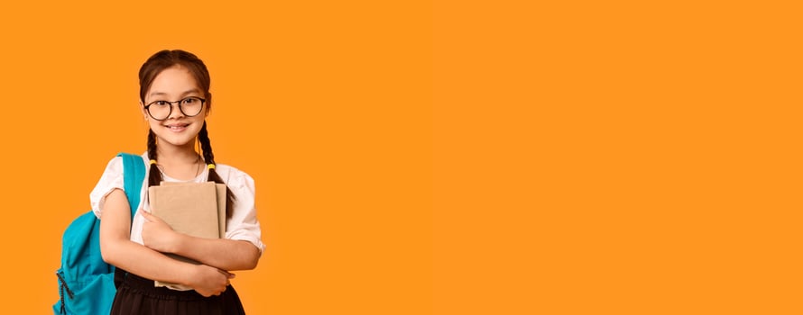 Back To School. Happy Asian Schoolgirl Embracing Book Standing With Backpack In Studio On Yellow Background. Free Space For Text photo