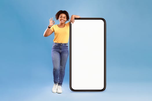 New awesome mobile app. Positive African American lady posing near huge cellphone with empty screen, showing okay gesture, recommending website, presenting space for advertisement on blue photo