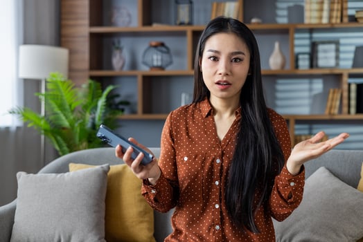 Asian woman with long hair in brown polka dot shirt holding phone looks confused. She sits on sofa at home expressing surprise. Modern interior visible in background. photo