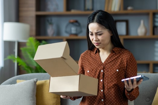 Asian woman holding phone, opening box with confused expression at home. Highlights online shopping mishap or delivery issue, showcasing customer dissatisfaction and unexpected packaging. photo