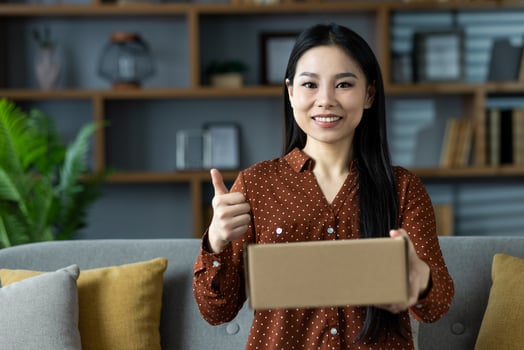 Asian woman with long hair and dotted shirt holding cardboard box, giving thumbs up on sofa. Captures happiness, success, home comfort, and positivity photo